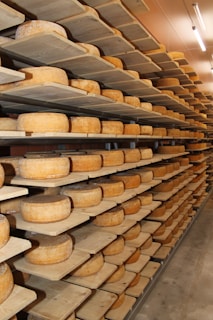 Rows of cheese wheels are neatly arranged on wooden shelves inside a storage or aging room. The shelves are stacked from floor to ceiling, and the room is well-lit with fluorescent lights.