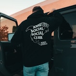 A person wearing a black hoodie with a graphic design of a map and text saying 'Anti Social Social Club North America' is standing near an open car door. The background features a building with brick walls and some greenery.