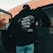 A person wearing a black hoodie with a graphic design of a map and text saying 'Anti Social Social Club North America' is standing near an open car door. The background features a building with brick walls and some greenery.