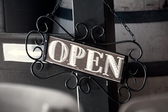 A vintage-style open sign hangs on a decorative wrought iron bracket. The background features wooden elements, including a barrel with metal bands. The sign displays the word 'OPEN' in bold, vintage lettering.
