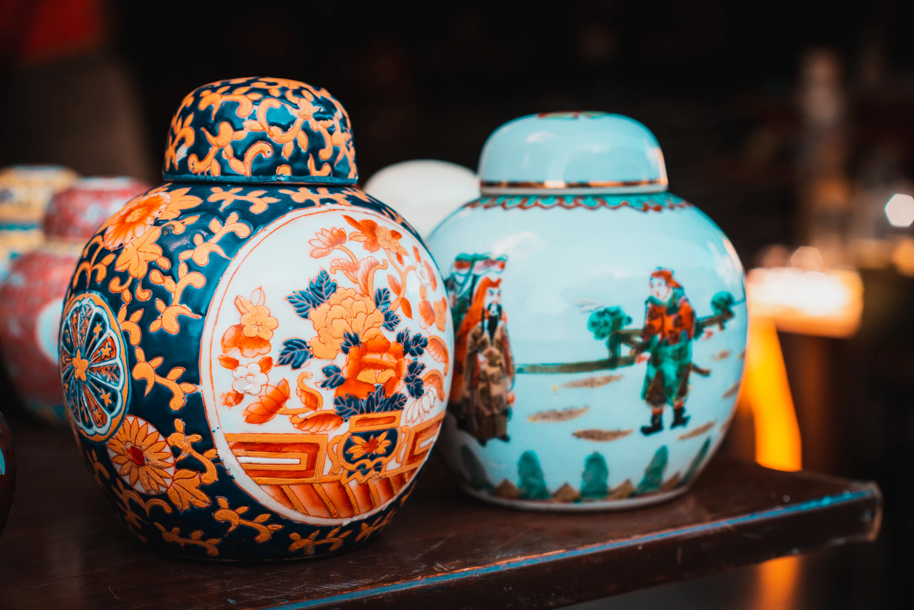 Two painted porcelain ginger jars, one orange and blue and one turquoise, standing on a dark shelf