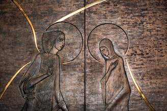 This artwork features two stylized figures in profile, possibly depicting a spiritual or religious scene. The figures are surrounded by halos and are carved into a textured metallic surface, with golden accents emphasizing their forms.