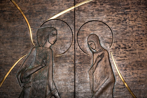 This artwork features two stylized figures in profile, possibly depicting a spiritual or religious scene. The figures are surrounded by halos and are carved into a textured metallic surface, with golden accents emphasizing their forms.