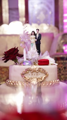 A decorative wedding cake features a topper with a figurine of a bride and groom. The cake is adorned with red and white flowers, gold detailing, and a personalized name plaque. The background shows a blurred, ornate setting with rich purple and gold hues.