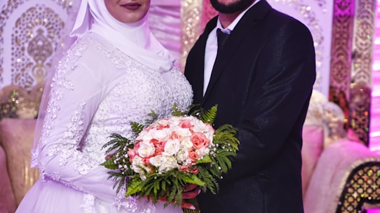 A couple in formal attire is standing in an ornately decorated room. The bride is wearing a white dress with intricate embroidery and a hijab, holding a bouquet of pink and white flowers with green foliage. The groom is in a black suit with a white shirt and black tie. The background features luxurious seating and elegant, patterned decorations in gold and cream tones.