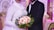 A couple in formal attire is standing in an ornately decorated room. The bride is wearing a white dress with intricate embroidery and a hijab, holding a bouquet of pink and white flowers with green foliage. The groom is in a black suit with a white shirt and black tie. The background features luxurious seating and elegant, patterned decorations in gold and cream tones.