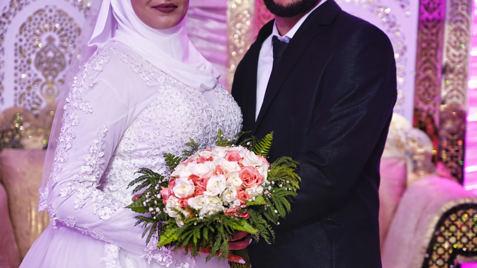 A radiant bride and groom in traditional Minang attire, standing beneath an ornate pelaminan adorned with deep gold and ivory fabrics.