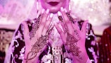 A close-up of a person's hands adorned with intricate henna designs. The henna features floral and geometric patterns, with one hand showing the name 'Sohail' incorporated into the design. The person is wearing an ornate outfit in rich fabric with embroidery, and there is a soft pink and purple lighting.
