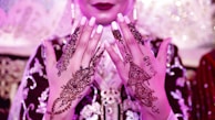 A close-up of a person's hands adorned with intricate henna designs. The henna features floral and geometric patterns, with one hand showing the name 'Sohail' incorporated into the design. The person is wearing an ornate outfit in rich fabric with embroidery, and there is a soft pink and purple lighting.