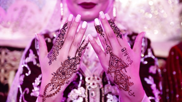 A close-up of a person's hands adorned with intricate henna designs. The henna features floral and geometric patterns, with one hand showing the name 'Sohail' incorporated into the design. The person is wearing an ornate outfit in rich fabric with embroidery, and there is a soft pink and purple lighting.