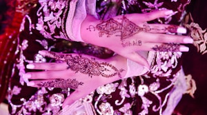Two hands adorned with intricate henna designs, featuring floral and geometric patterns. There is also a name inscribed within the henna on one of the hands. The hands are placed against a richly embroidered fabric in dark maroon and gold colors.