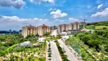 A wide road lined with lush green trees leads towards a group of multi-story apartment buildings. Behind these buildings, a hill covered with vegetation is visible. A construction crane stands next to a partially built structure on the right side of the image. The sky is partly cloudy, providing a bright and clear atmosphere to the scene.
