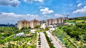 A wide road lined with lush green trees leads towards a group of multi-story apartment buildings. Behind these buildings, a hill covered with vegetation is visible. A construction crane stands next to a partially built structure on the right side of the image. The sky is partly cloudy, providing a bright and clear atmosphere to the scene.