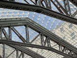 Intricate metal beams and trusses crisscross beneath a glass roof, creating geometric patterns. The transparent ceiling allows natural light to filter through, enhancing the modern architectural design. The steel framework exhibits industrial strength and complexity.
