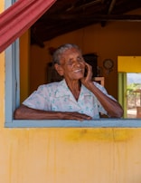 An elderly person with a warm smile is leaning on a window sill. The individual is wearing a light-colored shirt with a pattern. The background features the interior of a rustic house with warm yellow tones on the walls and a red curtain. A clock is visible in the background, adding to the cozy atmosphere.