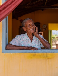 An elderly person with a warm smile is leaning on a window sill. The individual is wearing a light-colored shirt with a pattern. The background features the interior of a rustic house with warm yellow tones on the walls and a red curtain. A clock is visible in the background, adding to the cozy atmosphere.