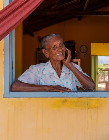 An elderly person with a warm smile is leaning on a window sill. The individual is wearing a light-colored shirt with a pattern. The background features the interior of a rustic house with warm yellow tones on the walls and a red curtain. A clock is visible in the background, adding to the cozy atmosphere.