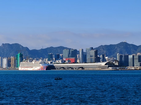A large cruise ship docked at a modern terminal, with a backdrop of a city skyline featuring numerous skyscrapers and a mountain range in the distance. The foreground shows a calm body of water with a small boat visible.