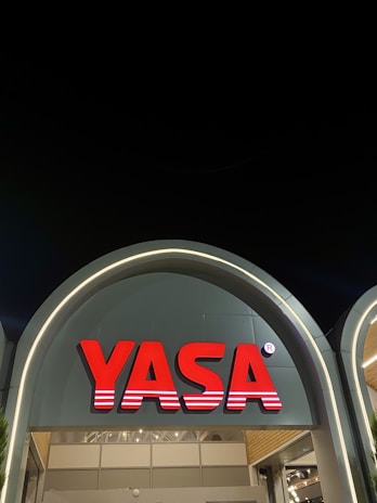 A modern architectural structure featuring an illuminated sign with the word 'YASA' in bold red letters. The sign is mounted on an arched facade that has a sleek, contemporary design with soft lighting accentuating its curves. The night sky provides a contrasting dark backdrop.