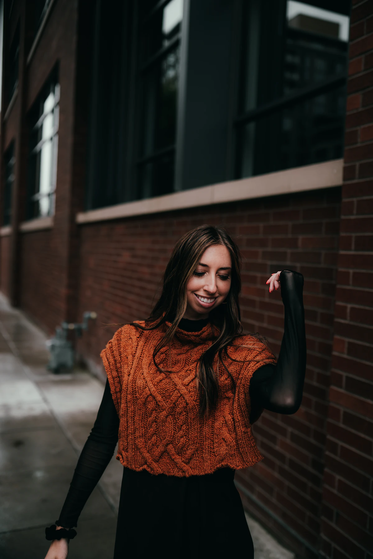 Woman in an orange sweater and black trousers by a brick wall