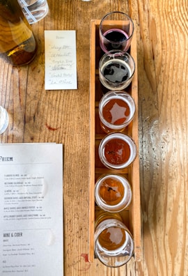 Five small glasses filled with different types of beer arranged in a wooden tray on a rustic wooden table. A menu and a handwritten note are placed beside the tray, listing the names of the beers. A bottle is visible at the top left corner.