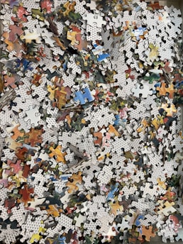 A large collection of jigsaw puzzle pieces spread out, showing a variety of colors and patterns. Each piece has black alphanumeric marks on the backside, and the front shows segments of a colorful image. The pieces are an assortment of shapes, indicating an unfinished puzzle.