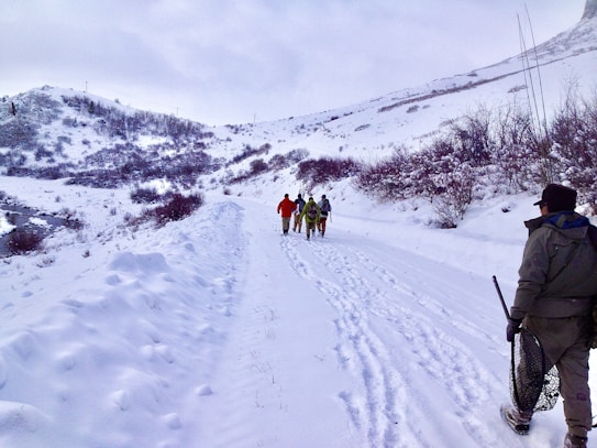 A snowy mountainous landscape with a group of people walking along a snow-covered path. The path is surrounded by snow-laden bushes and small hills. One person in the foreground is carrying a fishing net, suggesting a winter outdoor activity.