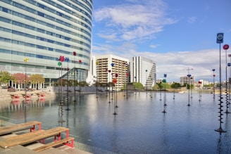 A modern cityscape with tall buildings, including a curved glass structure, surrounding a reflective pool. The pool is adorned with tall sculptures topped with colorful geometric shapes. Red benches line the edge of the pool alongside landscaped areas with small trees. The sky is clear with a few clouds, creating a bright and open atmosphere.