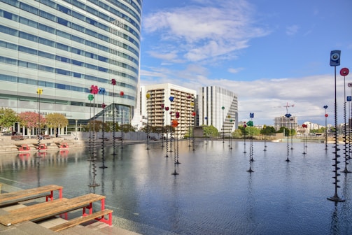 A modern cityscape with tall buildings, including a curved glass structure, surrounding a reflective pool. The pool is adorned with tall sculptures topped with colorful geometric shapes. Red benches line the edge of the pool alongside landscaped areas with small trees. The sky is clear with a few clouds, creating a bright and open atmosphere.