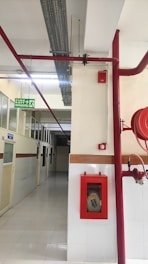 A corridor with tiled floor and walls partially lined with wood. Red fire safety equipment, including pipes and a fire hose cabinet, is mounted on the walls. Above, there are industrial ceiling fixtures and a green emergency exit sign.