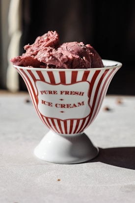A stylish white bowl with red vertical stripes and text reading 'Pure Fresh Ice Cream' contains a generous serving of pink ice cream. The setup is on a light gray surface with a dark blurred background, suggesting natural light casting shadows.