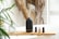 A black aroma diffuser emits a delicate stream of mist. It sits on a wooden board next to several small bottles of essential oils. A stylish glass soap dispenser with a golden pump is also on the board. Green leaves and dried plants in the background add a natural touch to the bright, tranquil setting.