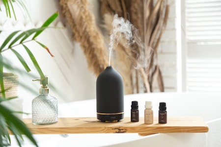 A black aroma diffuser emits a delicate stream of mist. It sits on a wooden board next to several small bottles of essential oils. A stylish glass soap dispenser with a golden pump is also on the board. Green leaves and dried plants in the background add a natural touch to the bright, tranquil setting.