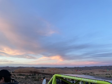 The photo captures a scenic desert landscape during sunset, with expansive skies painted in soft hues of blue, pink, and orange. Rolling hills and sparse vegetation can be seen in the background. A lime green vehicle with a visible website on its side is parked in the foreground, with a few people nearby.