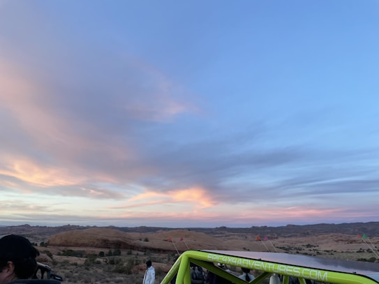 The photo captures a scenic desert landscape during sunset, with expansive skies painted in soft hues of blue, pink, and orange. Rolling hills and sparse vegetation can be seen in the background. A lime green vehicle with a visible website on its side is parked in the foreground, with a few people nearby.