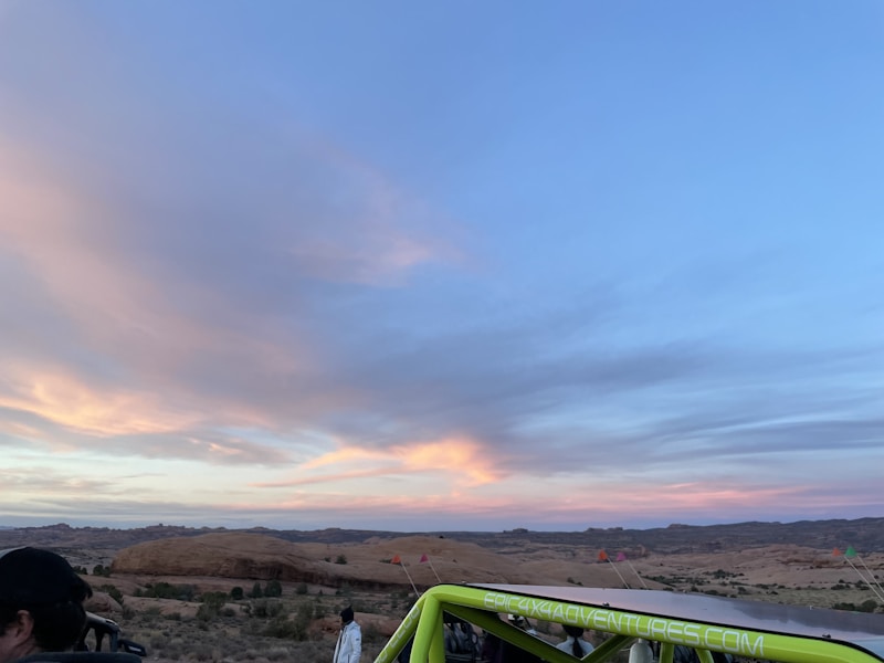 The photo captures a scenic desert landscape during sunset, with expansive skies painted in soft hues of blue, pink, and orange. Rolling hills and sparse vegetation can be seen in the background. A lime green vehicle with a visible website on its side is parked in the foreground, with a few people nearby.