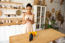 A woman in a light dress stands in a well-organized kitchen, holding a small black bottle with a bowl of oranges in front of her on a wooden table. The kitchen features white cabinets, open shelving with cups and jars, and a variety of hats and plants in the background, adding to the cozy and natural atmosphere.