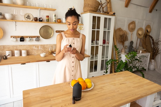 A woman in a light dress stands in a well-organized kitchen, holding a small black bottle with a bowl of oranges in front of her on a wooden table. The kitchen features white cabinets, open shelving with cups and jars, and a variety of hats and plants in the background, adding to the cozy and natural atmosphere.