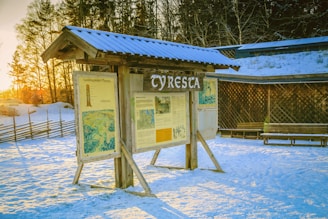 A wooden information board with a roof displays maps and information about the Tyresta area. It stands on snow-covered ground with sunlight casting long shadows. Trees and a rustic fence are in the background, with a wooden lattice wall and bench nearby.