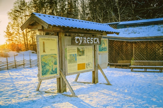 A wooden information board with a roof displays maps and information about the Tyresta area. It stands on snow-covered ground with sunlight casting long shadows. Trees and a rustic fence are in the background, with a wooden lattice wall and bench nearby.