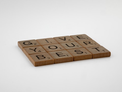 The image features wooden letter tiles arranged to spell 'GIVE YOUR BEST' on a white background. Each tile displays a letter with a small number at the bottom corner, resembling Scrabble tiles.