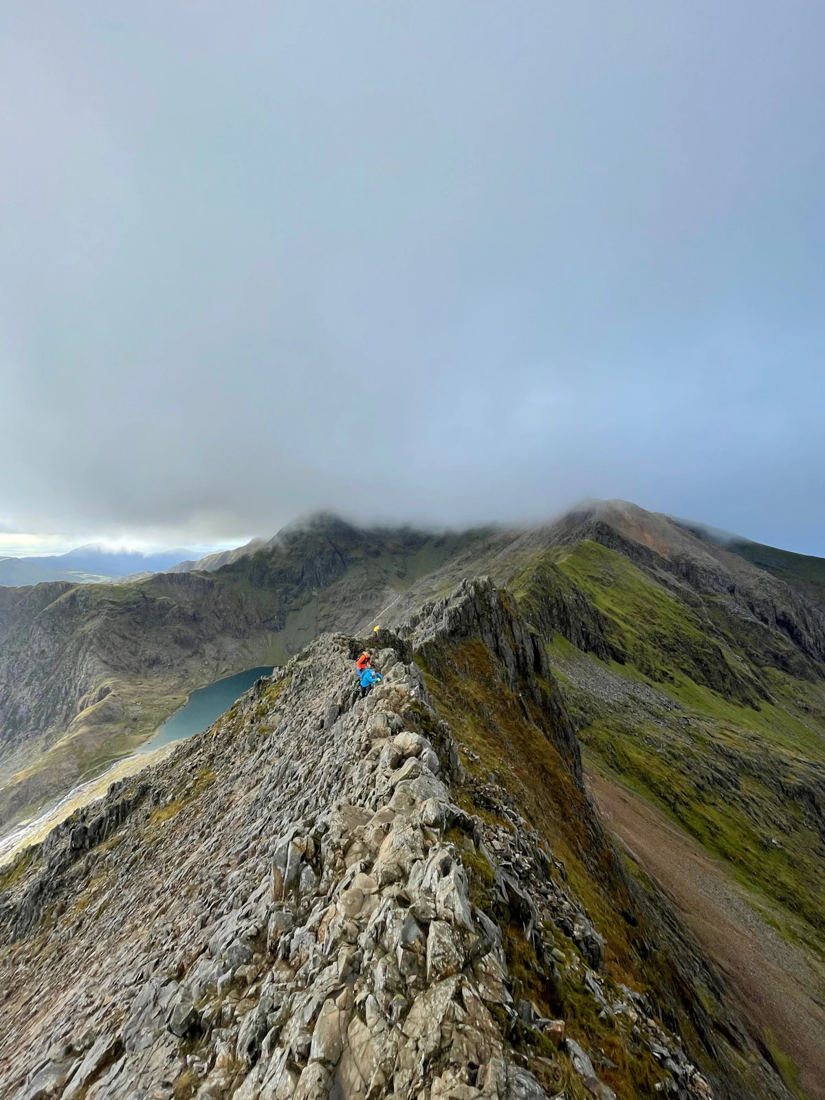 Climbers on the rocky crest of Crib Goch above a lake, with cloud low over Snowdon
