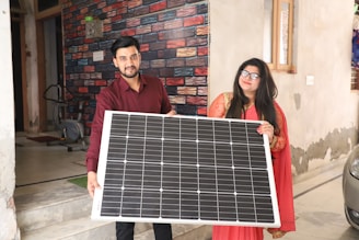 Two people are standing indoors, holding a large solar panel together. The person on the left is wearing a red shirt and the person on the right is in a red and gold traditional outfit. The background features a colorful brick-patterned wall and part of an exercise machine is visible.