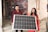 Two people are standing indoors, holding a large solar panel together. The person on the left is wearing a red shirt and the person on the right is in a red and gold traditional outfit. The background features a colorful brick-patterned wall and part of an exercise machine is visible.