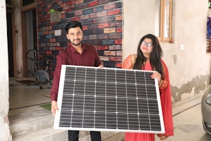 Two people are standing indoors, holding a large solar panel together. The person on the left is wearing a red shirt and the person on the right is in a red and gold traditional outfit. The background features a colorful brick-patterned wall and part of an exercise machine is visible.