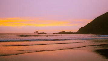 A serene beach at sunset with gentle waves lapping at the shore. The sky displays a gradient of warm colors from yellow to pink. Several surfers can be seen in the water, and a rocky promontory juts into the sea while a distant lighthouse is visible on the horizon.