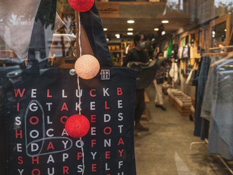 A close-up view of a tote bag with text in a shop window. The tote features a black design with red and white text interspersed in a grid. String lights with red and white spheres hang next to it. Inside the store, there are racks with clothing and a person wearing a mask, adding a bustling yet cozy atmosphere.