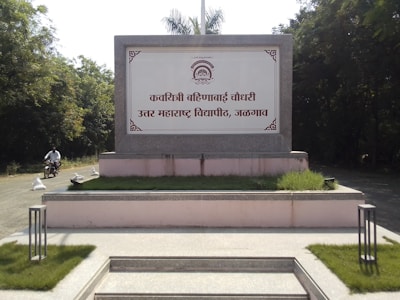 A large stone signboard displayed prominently, featuring Marathi text within an ornate border. The signboard is situated on raised ground surrounded by trimmed grass and pathway. On the left, a person is riding a motorcycle down a road that curves behind the sign. Dense green foliage frames the scene, suggesting a natural or suburban setting.