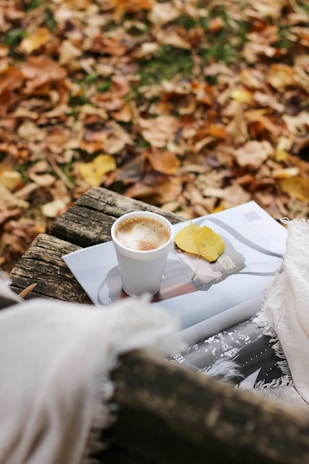 Close-up of a steaming cup of coffee resting on a wooden stump, surrounded by pinecones and autumn leaves.