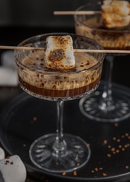 A close-up of a cocktail glass filled with a coffee-based beverage, topped with a roasted marshmallow skewer. The drink appears creamy and is garnished with cocoa or coffee powder, suggesting a dessert-like quality. The setting has a cozy, warm ambiance with a dark background that contrasts with the glass and marshmallow.